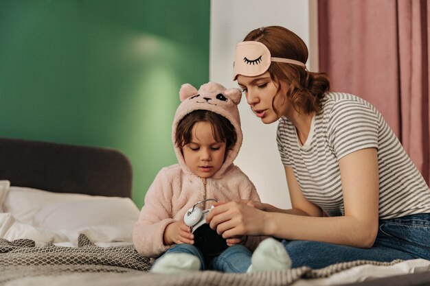 Mulher com máscara de dormir ensina a filha de pijama fofo como ligar o despertador.