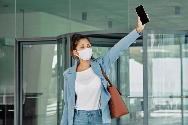 Mulher com máscara médica e smartphone chamando um táxi no aeroporto durante a pandemia