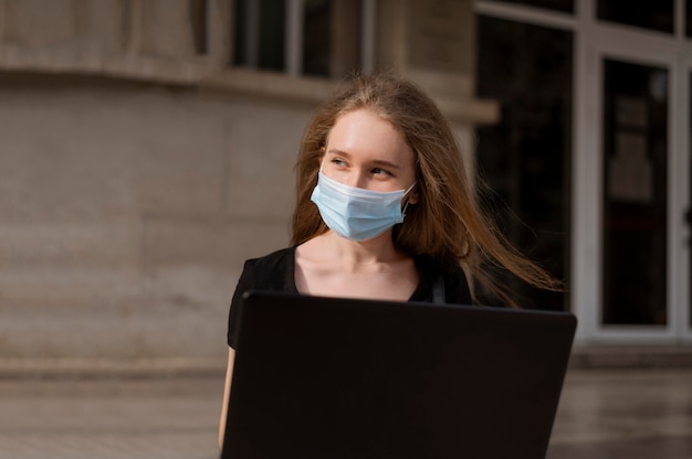 Mulher com máscara médica sentada na escada do lado de fora enquanto trabalha no laptop
