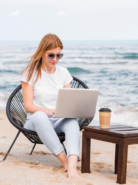 Mulher com óculos de sol na cadeira de praia, trabalhando no laptop