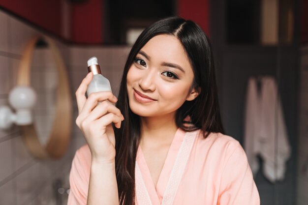 mulher com pele saudável é fofa sorrindo e posando com creme facial na parede do banheiro