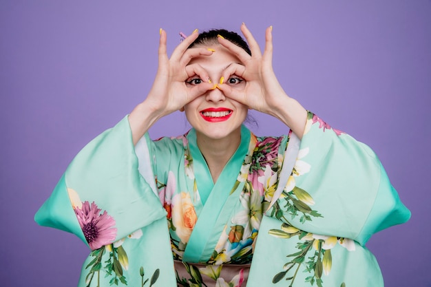 Mulher com quimono japonês tradicional fazendo gesto binocular com os dedos feliz e alegre no roxo