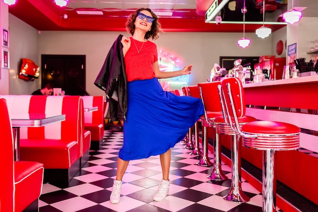 mulher com roupa colorida num café retro vintage dos anos 50 dançando com jaqueta, saia azul e camisa vermelha, óculos escuros se divertindo em clima alegre