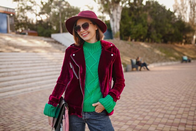 mulher com roupa da moda no estilo outono andando na rua usando uma jaqueta de veludo roxo, óculos escuros e chapéu