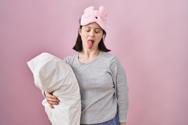 Mulher com síndrome de down usando máscara de dormir abraçando travesseiro enfiando a língua feliz com o conceito de emoção de expressão engraçada