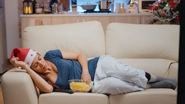 Mulher com sono e chapéu de papai noel assistindo televisão no sofá