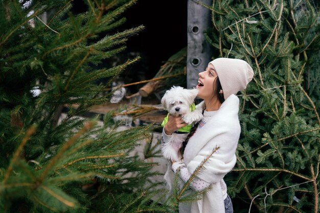 Mulher com um cachorro branco nos braços perto de uma árvore de Natal verde no mercado