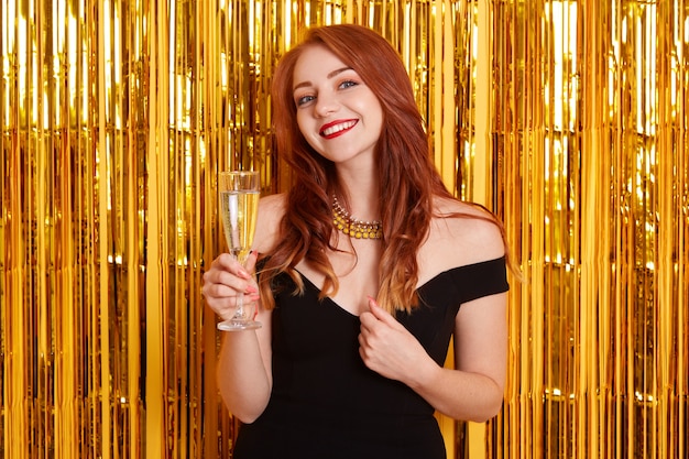 Mulher com um sorriso encantador, comemorando o ano novo, segurando uma taça de vinho, usando um elegante vestido preto, posando contra uma parede amarela com enfeites dourados.