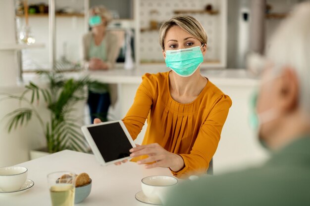 Mulher com uma máscara facial mostrando algo no touchpad para seu pai sênior em casa