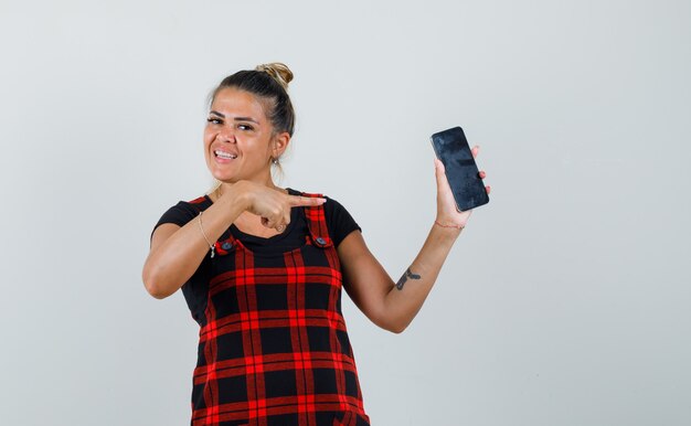 Mulher com vestido de avental, apontando para o celular e olhando orgulhosa, vista frontal.