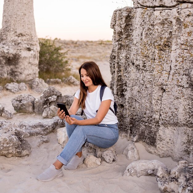 Mulher com vista frontal olhando para o telefone enquanto está sentada em uma pedra