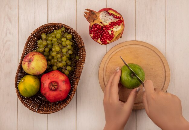 Mulher com vista superior corta maçã verde na tábua com maçãs e uvas com romãs na cesta na parede branca