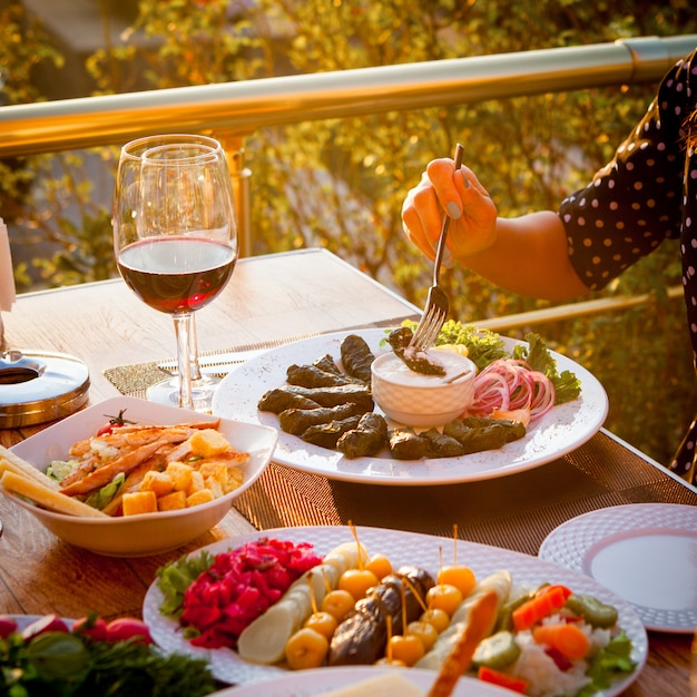 Mulher comendo folhas de uva recheada com diferentes tipos de saladas e um copo de vinho em uma mesa com árvores no fundo. vista de alto ângulo.