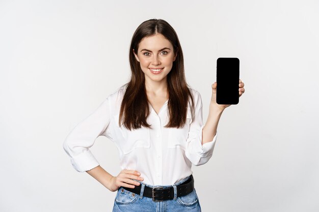 Mulher confiante em roupas corporativas, mostrando a tela do smartphone, interface móvel de um aplicativo, sobre fundo branco.