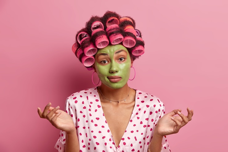 Foto grátis mulher confusa e inconsciente estende as mãos para os lados, enfrenta dilemas, aplica máscara facial verde para ficar bonita, usa modeladores de cabelo para um penteado perfeito, vestida com robe de seda, hesita sobre algo