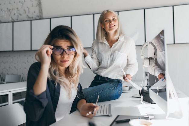 Mulher confusa na jaqueta preta, segurando os óculos, enquanto sua colega loira está sentada na mesa do escritório. Retrato interior da secretária triste, posando durante um duro dia de trabalho.