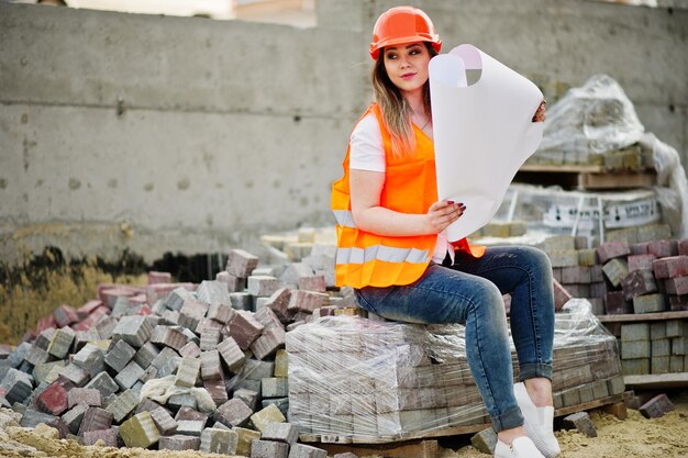 Mulher construtora de engenheira em colete uniforme e capacete protetor laranja segura papel de plano de layout de negócios sentado no pavimento