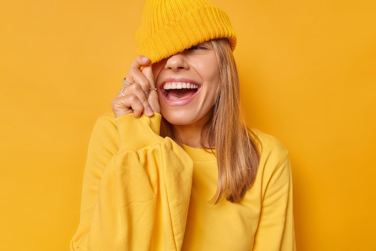 Foto grátis mulher contras olhos com chapéu tem expressão alegre vestida com jumper casual isolado em amarelo brilhante. adolescente despreocupada brincando com uma expressão engraçada