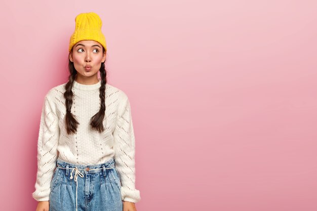 Mulher coreana bonita e terna mantém os lábios arredondados, pensa em algo, imagina uma cena em mente, vestida com roupas da moda, posa contra uma parede rosa