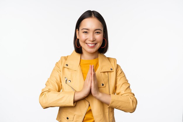 Mulher coreana feliz olhando esperançosa pedindo ajuda favor implorando de pé com gesto de namaste e sorrindo em pé sobre fundo branco