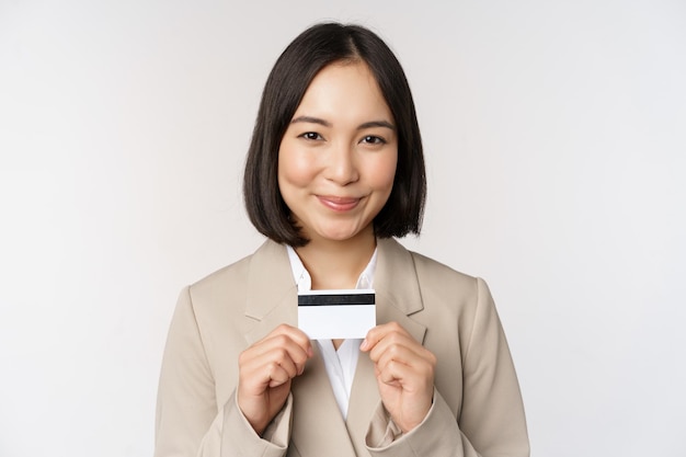 Mulher corporativa asiática sorridente de funcionário de escritório mostrando cartão de crédito em pé sobre fundo branco em terno bege