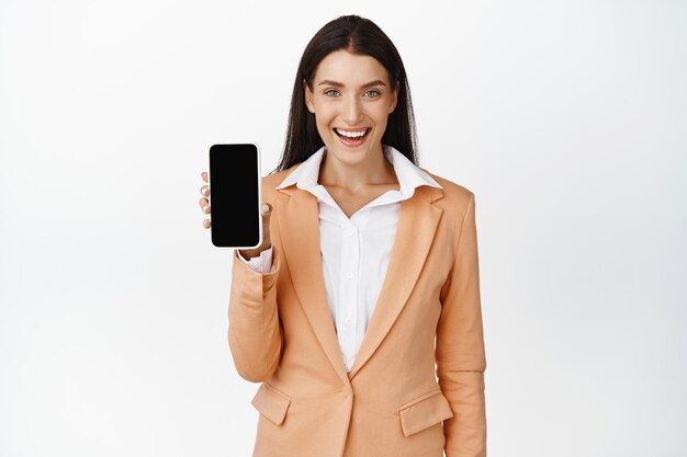 Mulher corporativa bem-sucedida, mostrando a tela do celular e sorrindo, recomendando o fundo branco do aplicativo para smartphone