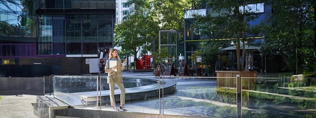 Foto grátis mulher corporativa elegante de terno em pé com laptop e documentos de trabalho usando telefone celular olhando