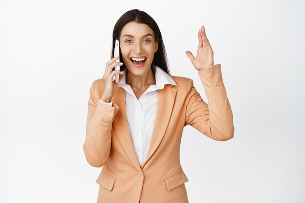 Mulher corporativa feliz recebe boas notícias no telefonema regozijando-se enquanto fala no smartphone em pé sobre fundo branco