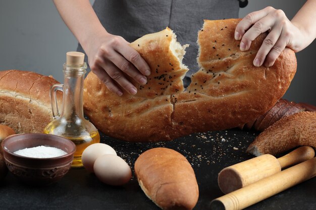 Mulher corta o pão ao meio na mesa escura com ovos, tigela de farinha e copo de óleo.