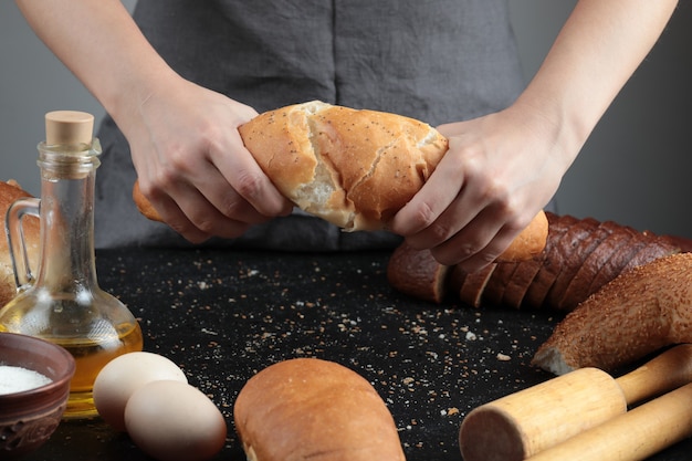 Mulher cortando o pão ao meio na mesa escura com ovos, tigela de farinha e copo de óleo.