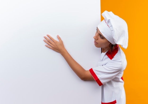 Mulher cozinheira satisfeita usando uniforme de chef, segurando e olhando para uma parede branca na parede amarela isolada com espaço de cópia