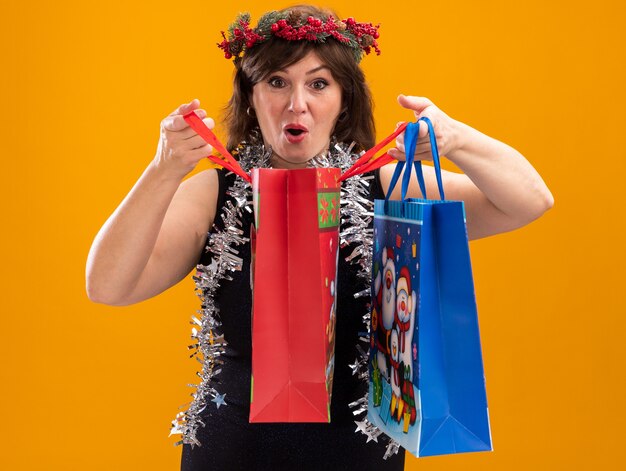 Mulher curiosa de meia-idade usando coroa de flores de Natal e guirlanda de ouropel em volta do pescoço segurando sacolas de presente de Natal, abrindo uma delas olhando para a câmera isolada em fundo laranja