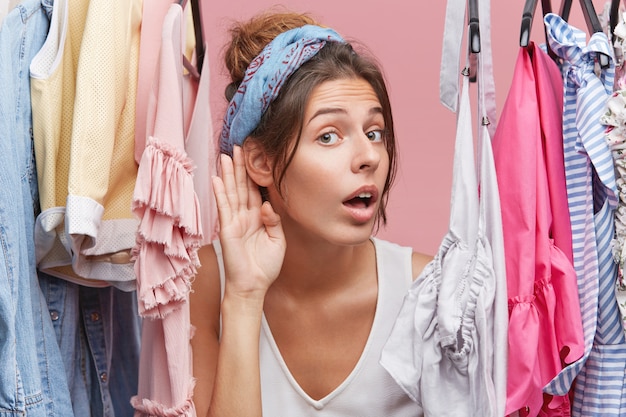 Mulher curiosa em pé no vestiário, tentando novas roupas coloridas, escutando o que as pessoas falam no próximo quarto. Mulher curiosa, mantendo a mão perto da orelha, ouvindo algo com atenção