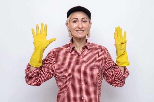 Mulher da limpeza com camisa xadrez e boné usando luvas de borracha e sorrindo alegremente mostrando o número nove com os dedos