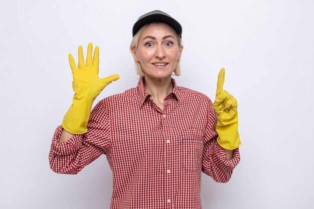 Mulher da limpeza com camisa xadrez e boné usando luvas de borracha, olhando para a câmera, sorrindo alegremente, mostrando o número seis com os dedos em pé sobre um fundo branco