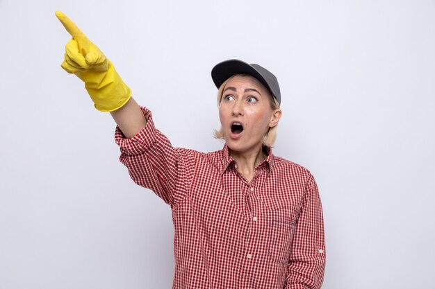 Mulher da limpeza com camisa xadrez e boné usando luvas de borracha, olhando para cima espantada e surpresa, apontando algo com o dedo indicador