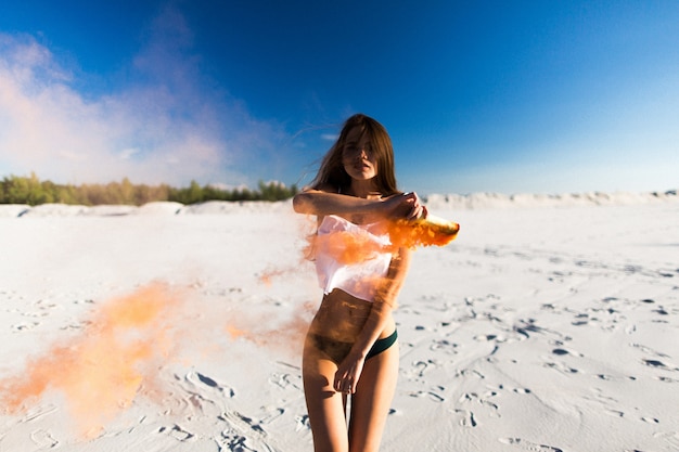 Mulher dança com fumaça de laranja na praia branca sob o céu azul