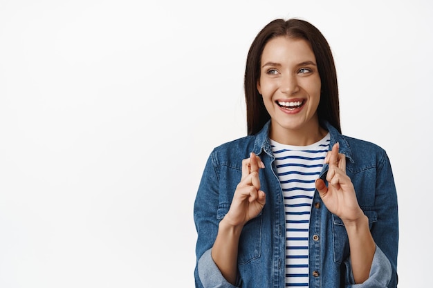 Mulher de 30 anos esperançosa, mulher adulta olha de lado para o logotipo, banner de venda e dedos cruzados, antecipando, esperando ganhar, sorrindo animado, de pé contra um fundo branco.