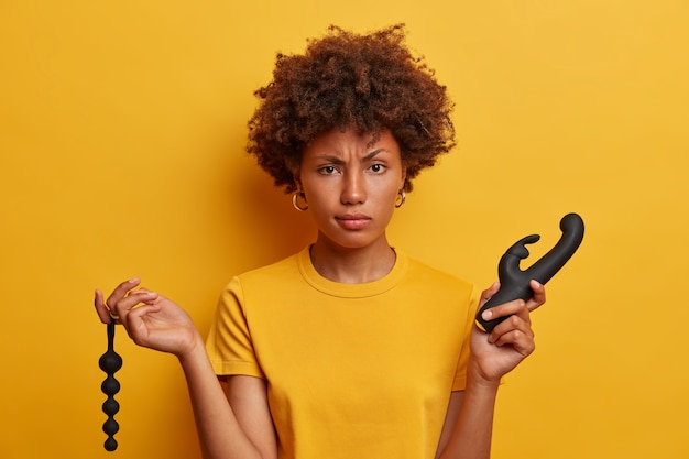 Mulher de aparência séria e descontente com cabelo afro escolhe entre contas anais e vibrador em sex shop, precisa de ferramenta para estimulação do clitóris e ponto G, penetração prazerosa. Orgasmo e prazer