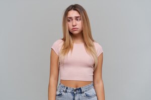 Mulher de aparência triste, garota desapontada com longos cabelos loiros. vestindo jeans e camiseta rosa. conceito de pessoas e emoção. observando à esquerda no espaço da cópia, isolado sobre a parede cinza