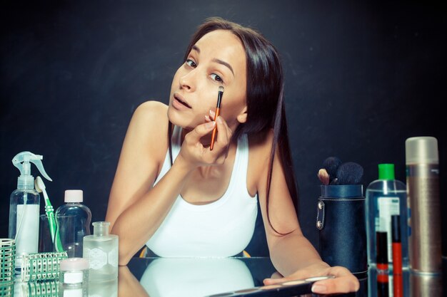 Mulher de beleza aplicando maquiagem. Linda garota olhando no espelho e aplicando cosméticos com um pincel. Bom dia, maquiagem e conceito de emoções humanas. Modelo caucasiana em estúdio