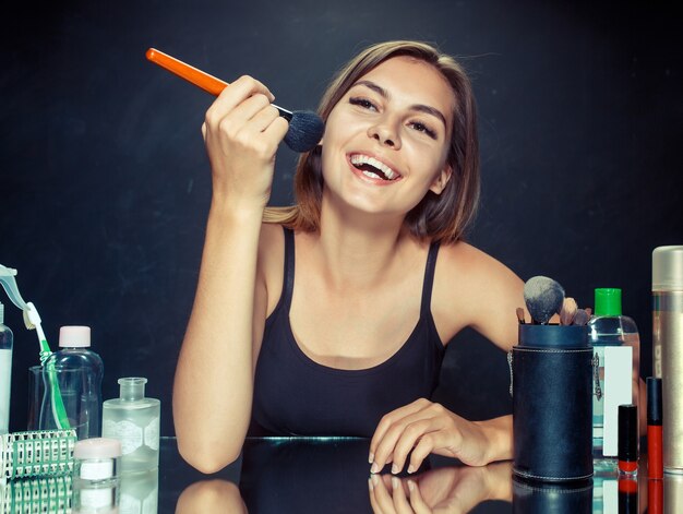 Mulher de beleza aplicando maquiagem. Linda garota olhando no espelho e aplicando cosméticos com um pincel grande. Bom dia, maquiagem e conceito de emoções humanas. Modelo caucasiana em estúdio