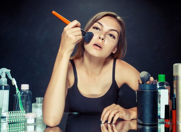 Mulher de beleza aplicando maquiagem. linda garota olhando no espelho e aplicando cosméticos com um pincel grande. bom dia, maquiagem e conceito de emoções humanas.