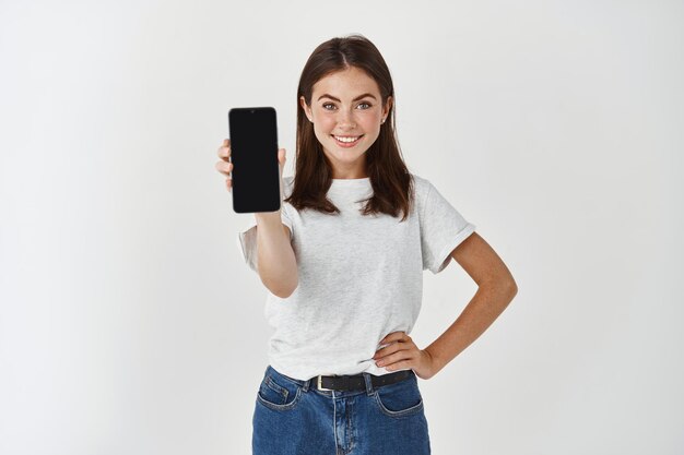 Mulher de beleza jovem segurando o celular de tela em branco e sorrindo, mostrando o visor do smartphone, em pé sobre uma parede branca.