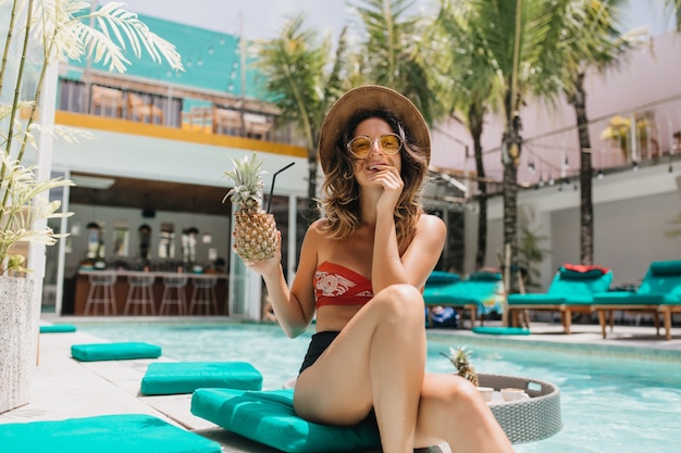 Mulher de biquíni, desfrutando de um coquetel de abacaxi no resort. mulher morena glamorosa posando com expressão de rosto interessado na piscina em dia de verão.