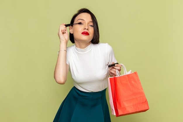 Mulher de blusa branca e saia verde segurando pacotes de compras e fazendo maquiagem