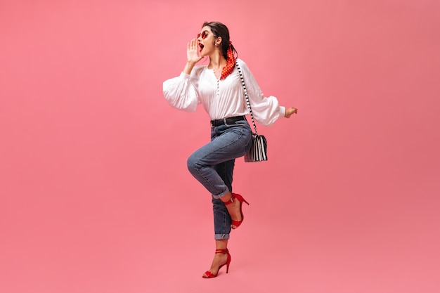 Mulher de bom humor grita no fundo rosa. menina elegante em blusa branca, jeans e salto alto, posando com bolsa listrada.