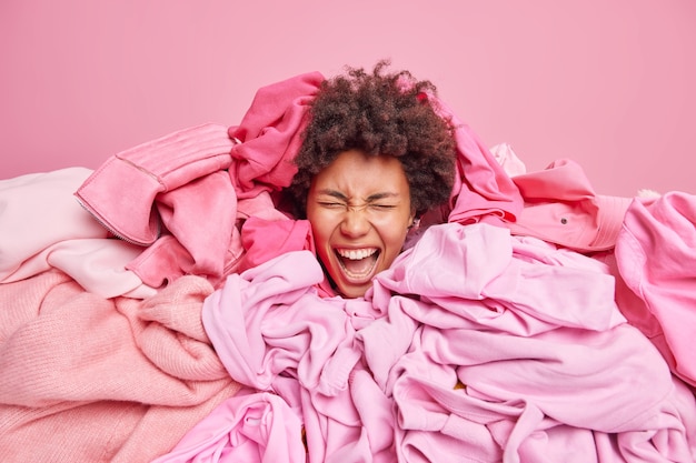 Mulher de cabelo encaracolado emocional cercada por uma pilha de roupas bagunçadas do armário exclama em voz alta mantém a boca aberta tem verdadeiro caos em casa ocupada lavando roupa. Tudo na cor rosa. Conceito de roupa