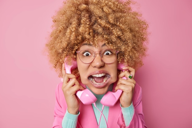 Mulher de cabelo encaracolado fala via telefone fixo segura aparelhos tris para estar em contato encara olhos esbugalhados e usa expansor de boca vestido com roupas formais isoladas em fundo rosa discute as últimas notícias