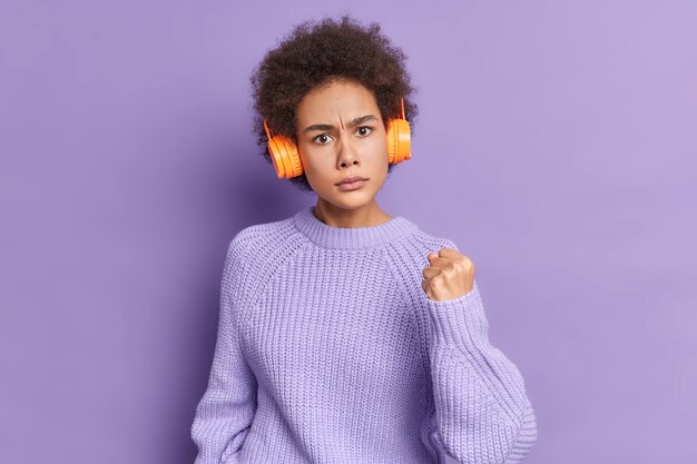 Mulher de cabelo encaracolado irritada mostra o punho, expressa raiva, ouve música com fones de ouvido sem fio